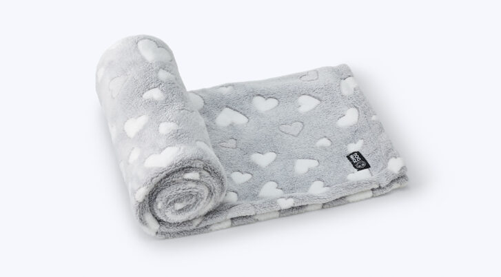 Szary, pluszowy koc dla psa w białe serca leży na białym tle i jest w części zwinięty w rulon, a pozostała część jest rozłożona i ma na sobie czarną naszywkę z logo John Doga.