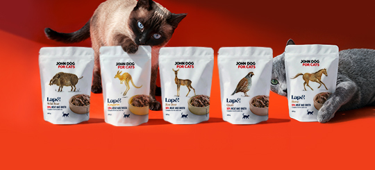 W centralnej części stoi pięć saszetek mokrej karmy dla kota Lape John Dog for Cats, jedna obok drugiej w rzędzie, w smakach dzik, kangur, sarna, przepiórka i konina. Kot rasy syjamskiej kładzie łapę na saszetce o smaku kangura. Na etykietach umieszczone są rysunki zwierząt na środku, a etykiety są w białym kolorze. Po prawej stronie kot rasy brytyjskiej obejmuje łapą saszetkę o smaku koniny. Tło obrazka jest w odcieniach barwy czerwonej.