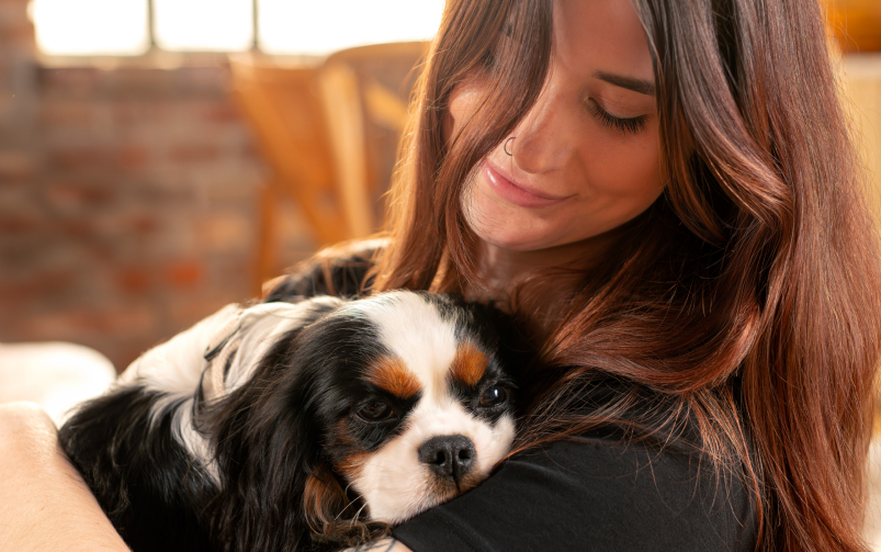 Kobieta przytula małego psa, kt&oacute;ry opiera głowę na jej ramieniu, a ona uśmiecha się.
