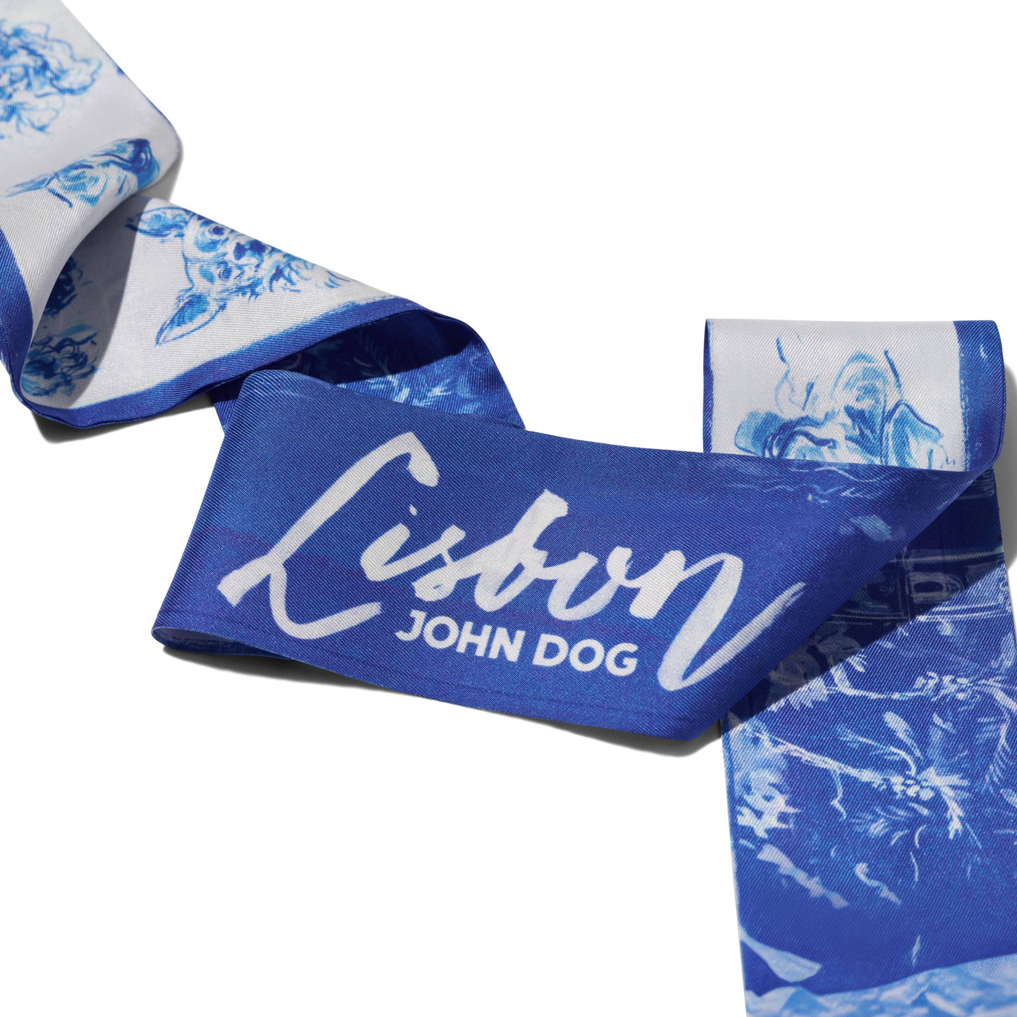 Zbliżenie na cienką, dwustronną apaszkę John Dog z kolekcji Lizbona w kolorze biało-granatowym ułożoną na kształt zygzaka. Z jednej strony są na niej rysunki granatowych portretów psów różnych ras na białym tle, a z drugiej rysunek białej panoramy miasta na granatowym tle i napis Lisbon w centrum.