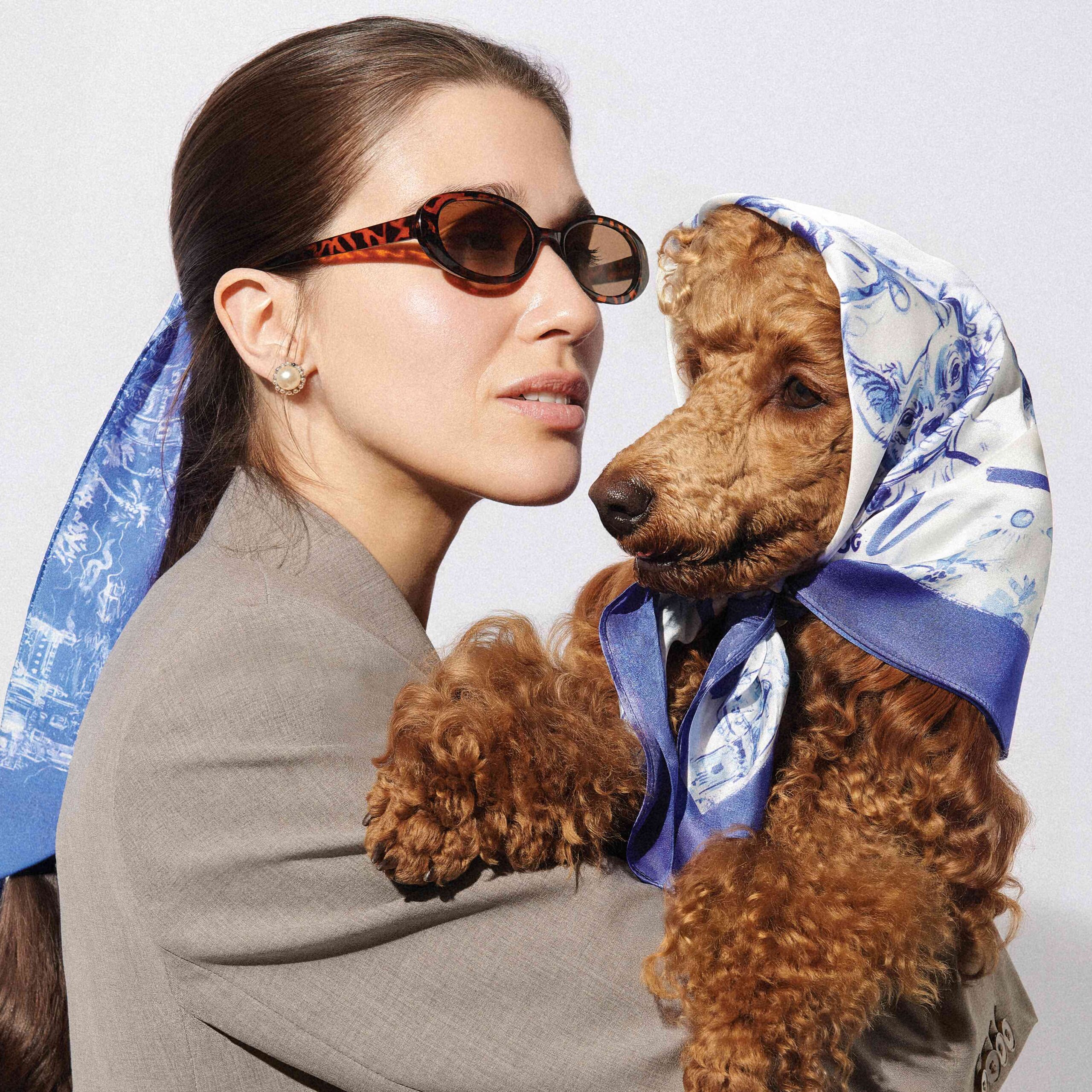 Modelka ubrana w szarą marynarkę, trzyma na rękach psa rasy pudel średni w kolorze płowym stojąc tyłem. Pies ma na sobie apaszkę John Dog z kolekcji Lizbona w kolorze błękitnym, modelka ma związane włosy w kitkę cienką apaszką John Dog z kolekcji Lizbona w kolorze błękitnym.