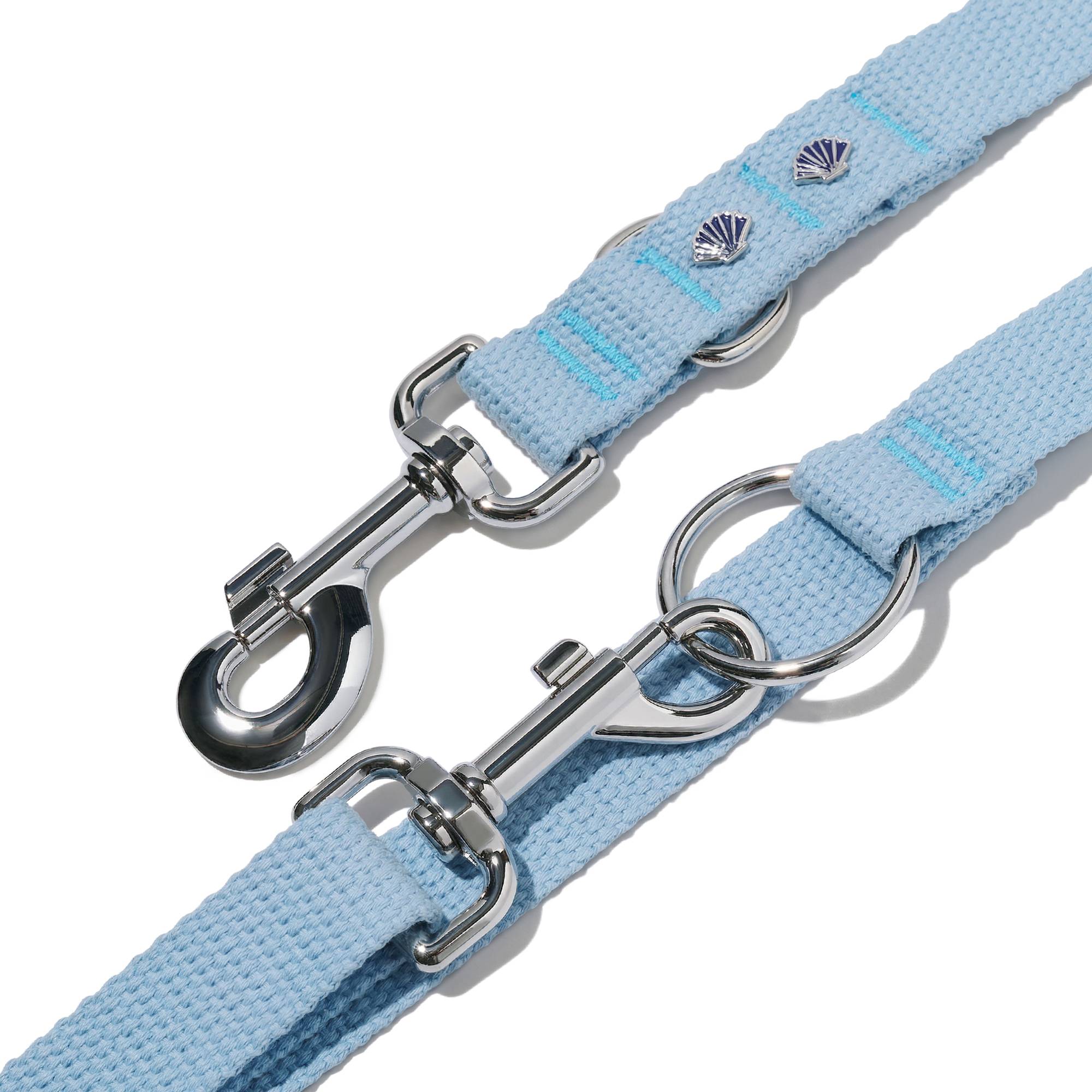Zbliżenie na zapięcie smyczy przepinanej John Dog z kolekcji Lizbona w błękitnym kolorze. Nad nim znajduje się taśma w kolorze błękitnym z dwoma pinami w kształcie muszelki i karabińczykiem.