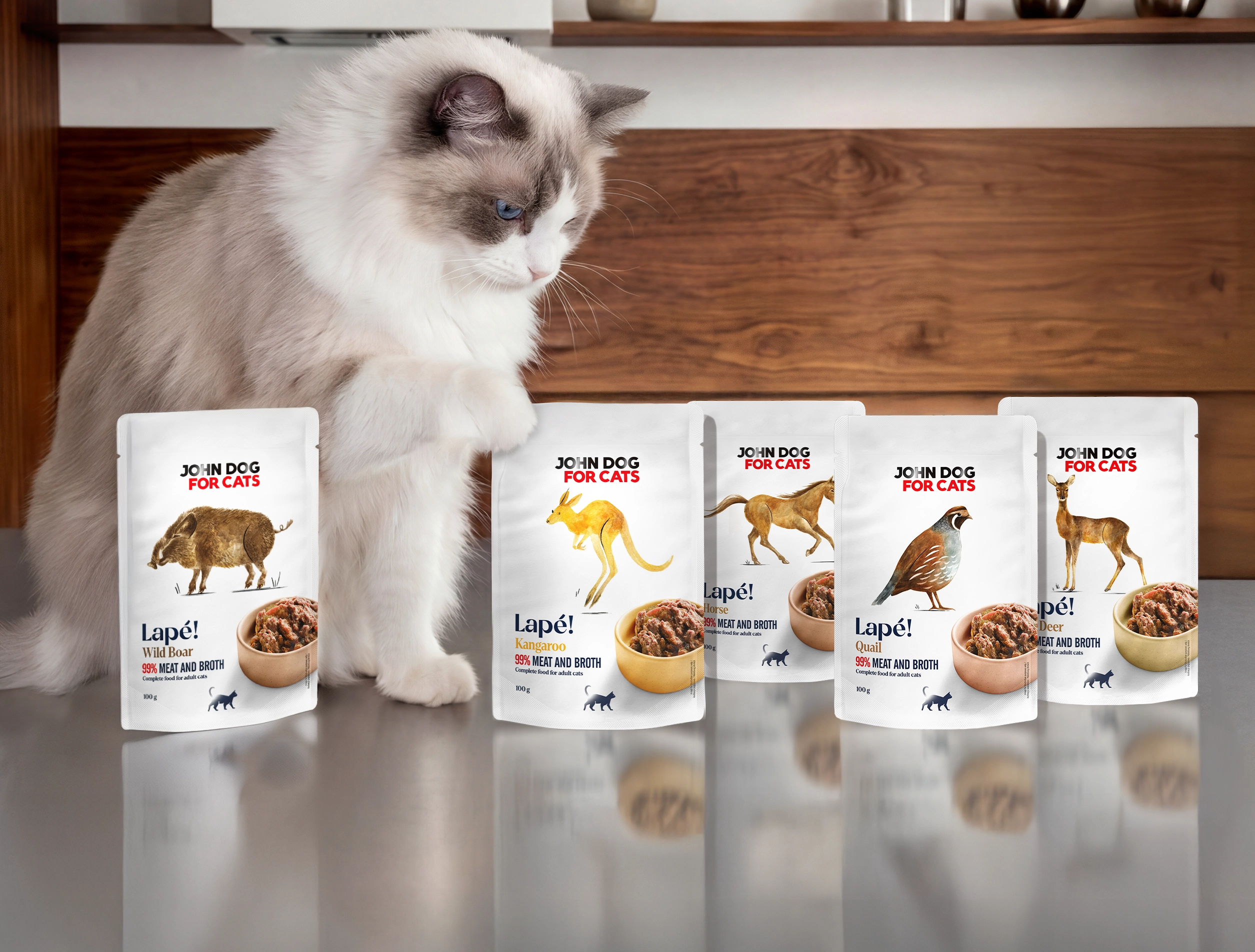 John Dog for Cats Lapé! – mokra karma dla kotów w saszetkach o różnych smakach. Na blacie stoi kilka opakowań karmy, obok których siedzi biało-szary kot i przygląda się produktom.