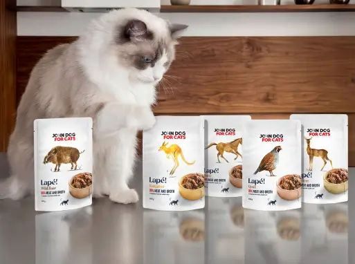 John Dog for Cats Lapé! – mokra karma dla kotów w saszetkach o różnych smakach. Na blacie stoi kilka opakowań karmy, obok których siedzi biało-szary kot i przygląda się produktom.
